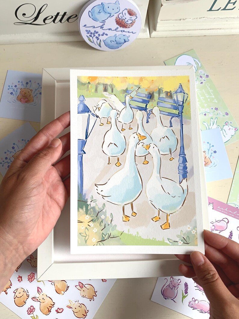 May include: A watercolor print featuring a group of white geese in a park setting. The artwork includes a blue bench, a lamppost, and yellow and green foliage. The print is held in a white frame, with additional stationery items visible.