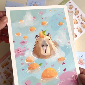 Op de afbeelding: Een grillige kunstprint met een capibara die ontspant in een bad met drijvende sinaasappels en roze bloemen. De capibara heeft een sinaasappel met een groen blad op zijn hoofd. De achtergrond is lichtblauw met een aquareleffect. Handtekening van de kunstenaar rechtsonder.