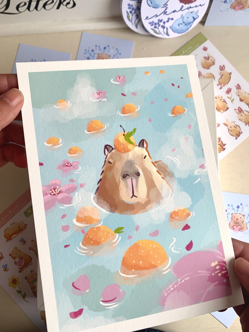 Op de afbeelding: Een grillige kunstprint met een capibara die ontspant in een bad met drijvende sinaasappels en roze bloemen. De capibara heeft een sinaasappel met een groen blad op zijn hoofd. De achtergrond is lichtblauw met een aquareleffect. Handtekening van de kunstenaar rechtsonder.