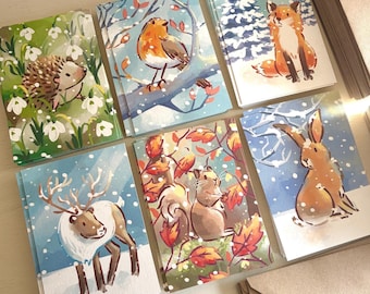 Op de afbeelding: Een verzameling van zes rechthoekige wenskaarten met aquarelillustraties van een egel, roodborstje, vos, rendier, eekhoorn en haas, elk afgebeeld tegen seizoensgebonden achtergronden met vallende sneeuw en bladeren.
