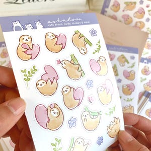 May include: A sheet of stickers featuring cute sloth illustrations. The sloths are depicted in various poses, including holding hearts, hanging from branches, and sleeping. The stickers are printed on white paper with a light blue background. The text "eshalou" and "cute prints, cards, stickers & more" is printed on the sheet.