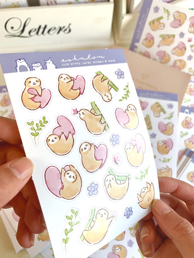 May include: A sheet of stickers featuring cute sloth illustrations. The sloths are depicted in various poses, including holding hearts, hanging from branches, and sleeping. The stickers are printed on white paper with a light blue background. The text "eshalou" and "cute prints, cards, stickers & more" is printed on the sheet.