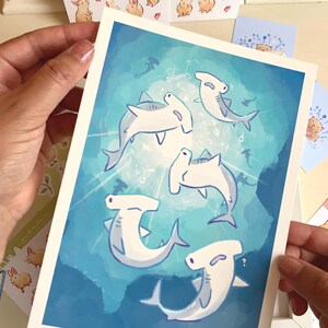 May include: A print featuring cartoon hammerhead sharks swimming in a blue ocean. The sharks are white with gray accents and are surrounded by bubbles and other small fish. Other cards with bunny and bear illustrations are also visible.