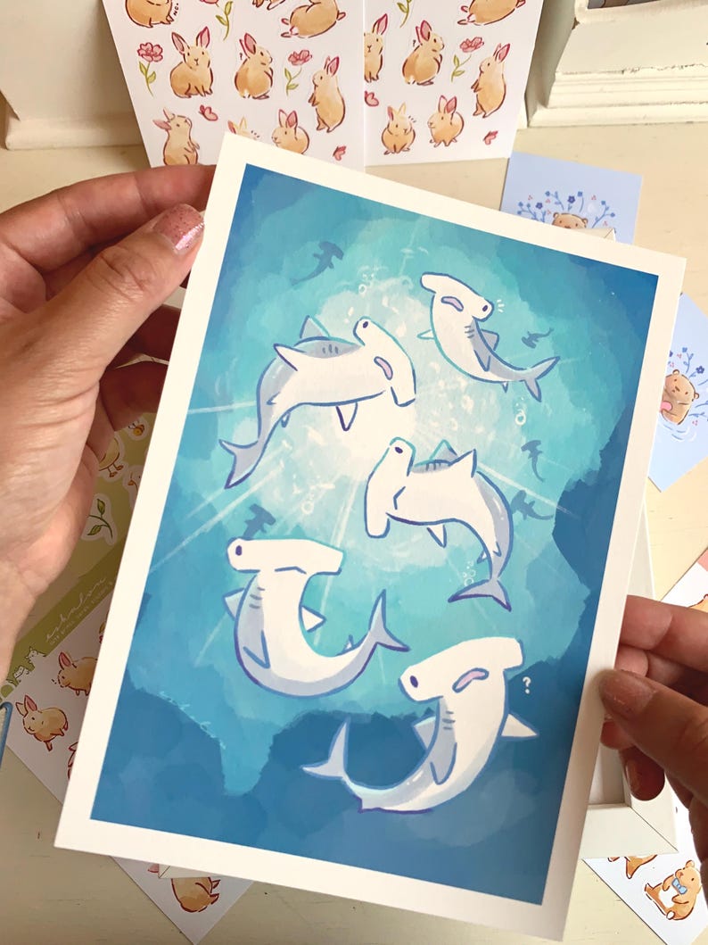 May include: A print featuring cartoon hammerhead sharks swimming in a blue ocean. The sharks are white with gray accents and are surrounded by bubbles and other small fish. Other cards with bunny and bear illustrations are also visible.