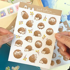 May include: A sheet of stickers featuring cute illustrations of hedgehogs in various poses. The stickers are on a white background with brown, yellow, and red accents. The text "eshalon cute prints, cards, stickers & more!" is printed in the top right corner.