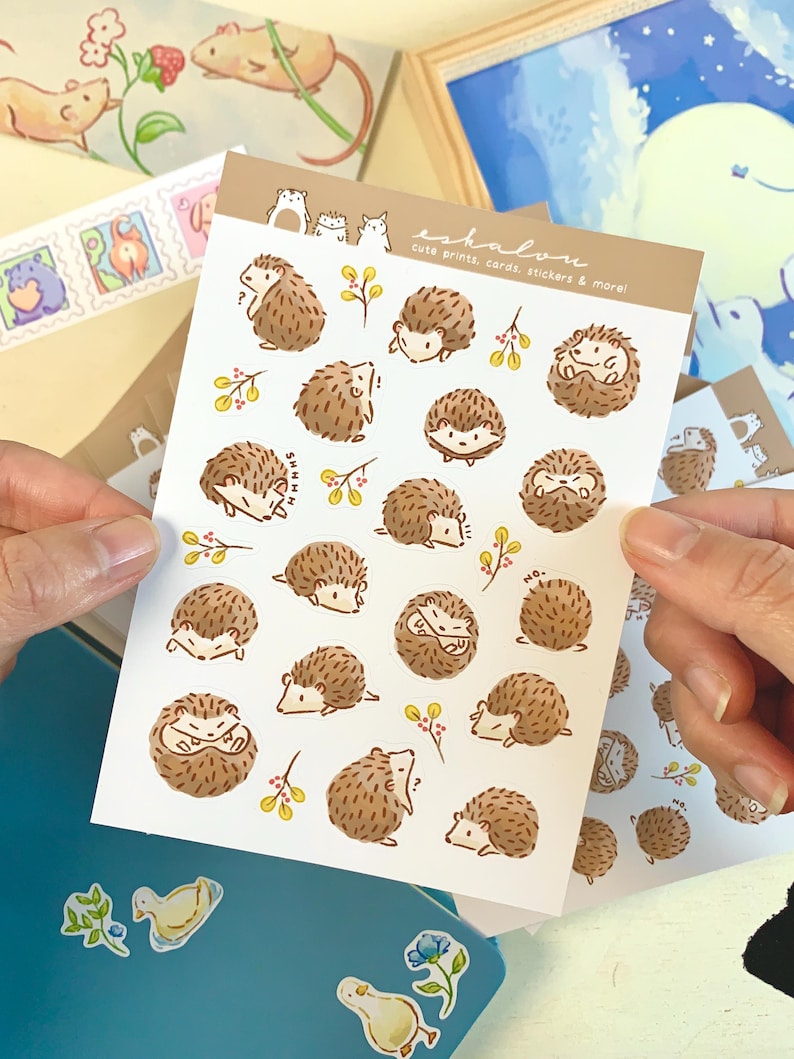 May include: A sheet of stickers featuring cute illustrations of hedgehogs in various poses. The stickers are on a white background with brown, yellow, and red accents. The text "eshalon cute prints, cards, stickers & more!" is printed in the top right corner.
