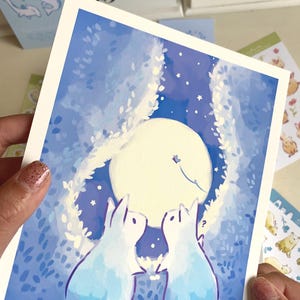 Op de afbeelding: Een aquarelprint met twee konijnen die naar een volle maan in een nachthemel kijken. Het kunstwerk is overwegend blauw en wit, met een grillige stijl. De print wordt door een persoon vastgehouden.