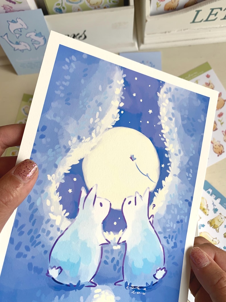Op de afbeelding: Een aquarelprint met twee konijnen die naar een volle maan in een nachthemel kijken. Het kunstwerk is overwegend blauw en wit, met een grillige stijl. De print wordt door een persoon vastgehouden.