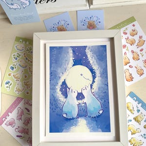 Op de afbeelding: Een ingelijste kunstdruk met twee lichtblauwe konijnen die naar een grote maan in een nachthemel kijken. Rondom het frame liggen diverse stickers met dierenillustraties, waaronder eenden, beren en konijnen, in zachte pastelkleuren.