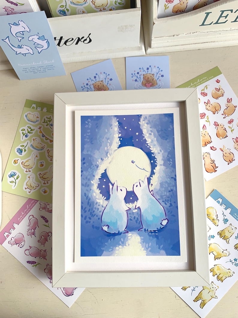 Op de afbeelding: Een ingelijste kunstdruk met twee lichtblauwe konijnen die naar een grote maan in een nachthemel kijken. Rondom het frame liggen diverse stickers met dierenillustraties, waaronder eenden, beren en konijnen, in zachte pastelkleuren.