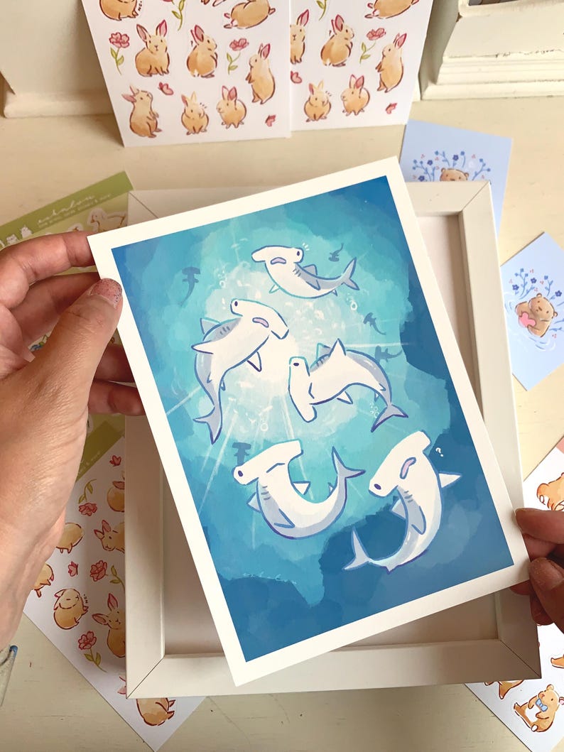 May include: A framed art print featuring a whimsical illustration of hammerhead sharks swimming in a blue ocean. The artwork has a bright, cartoonish style. Other cards and stickers with animal illustrations are visible.