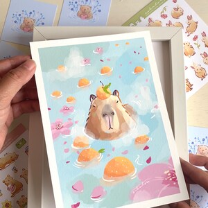 Op de afbeelding: Een kunstafdruk met een capibara die ontspant in een bad met sinaasappels en bloemblaadjes. De illustratie heeft een lichtblauwe achtergrond met wolken. Andere stickers en kaarten zijn zichtbaar, met een witte lijst om de afdruk.