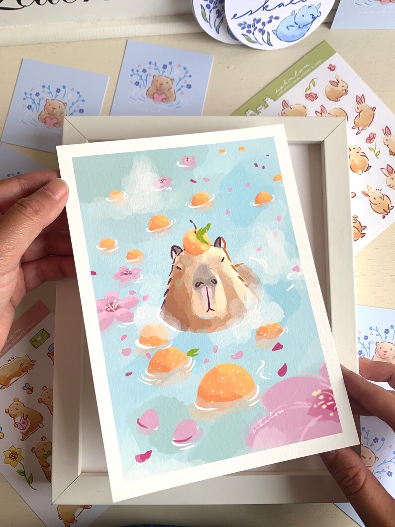 Op de afbeelding: Een kunstafdruk met een capibara die ontspant in een bad met sinaasappels en bloemblaadjes. De illustratie heeft een lichtblauwe achtergrond met wolken. Andere stickers en kaarten zijn zichtbaar, met een witte lijst om de afdruk.