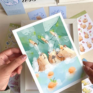 May include: A framed art print showing three capybaras enjoying a hot spring, with water cascading from bamboo pipes. The artwork uses a gentle palette of blues, greens, and oranges. Various stickers and cards with animal illustrations are scattered around the print.