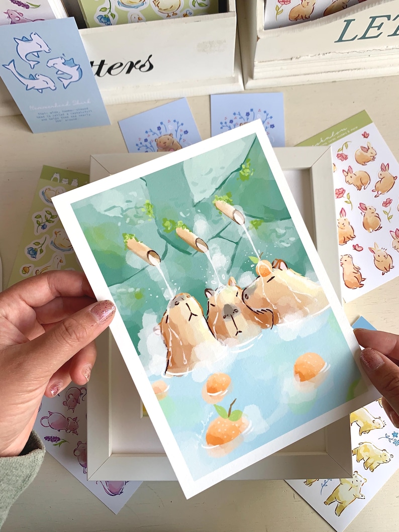 May include: A framed art print showing three capybaras enjoying a hot spring, with water cascading from bamboo pipes. The artwork uses a gentle palette of blues, greens, and oranges. Various stickers and cards with animal illustrations are scattered around the print.