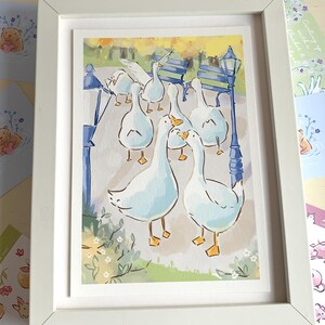 May include: A framed watercolor print featuring a group of white geese in a park setting. The artwork includes blue lampposts, benches, and greenery, all within a white frame. Additional small animal illustrations are visible around the frame.