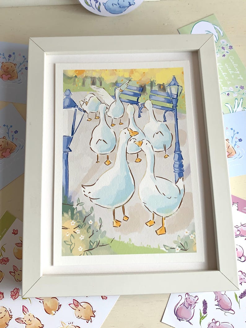 May include: A framed watercolor print featuring a group of white geese in a park setting. The artwork includes blue lampposts, benches, and greenery, all within a white frame. Additional small animal illustrations are visible around the frame.
