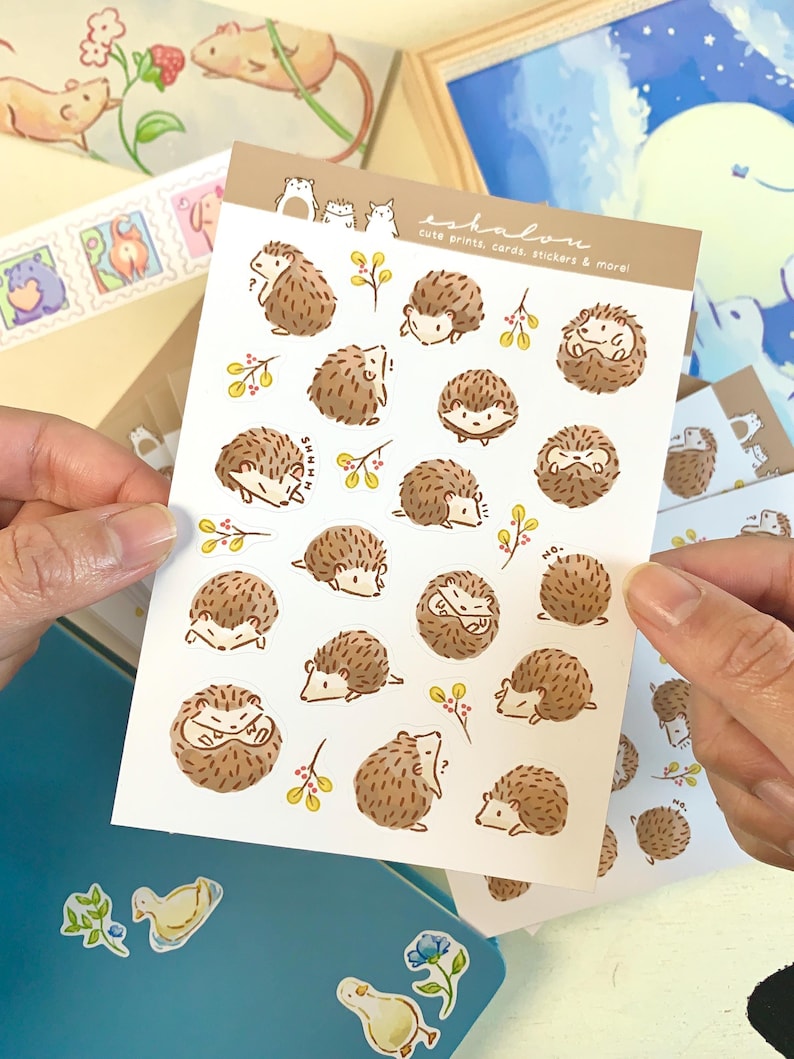 May include: A sheet of stickers featuring cute illustrations of hedgehogs in various poses. The stickers are on a white background with a brown border. The text "eshalawe cute prints, cards, stickers & more!" is printed at the top of the sheet.