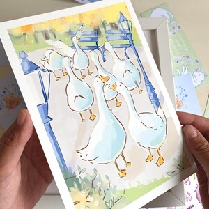 May include: A watercolor print featuring a flock of white geese in a park setting with a blue lamppost and bench. The artwork has a soft, pastel color palette. Other stationery items with animal illustrations are visible.