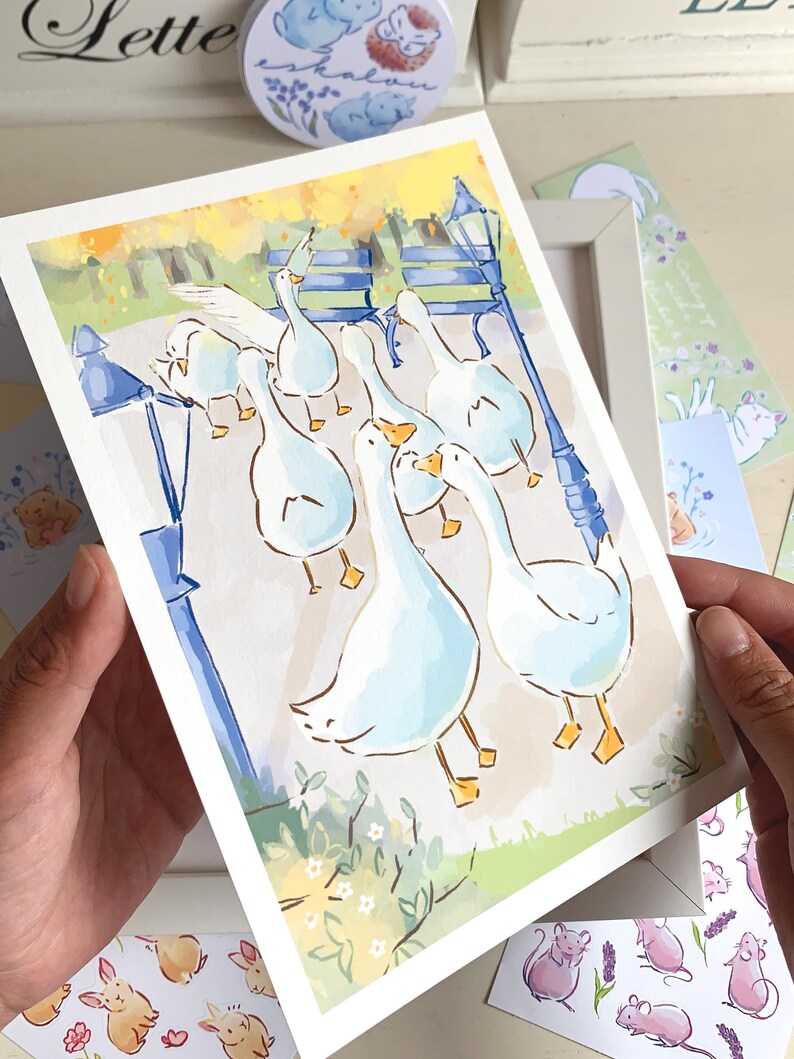 May include: A watercolor print featuring a flock of white geese in a park setting with a blue lamppost and bench. The artwork has a soft, pastel color palette. Other stationery items with animal illustrations are visible.