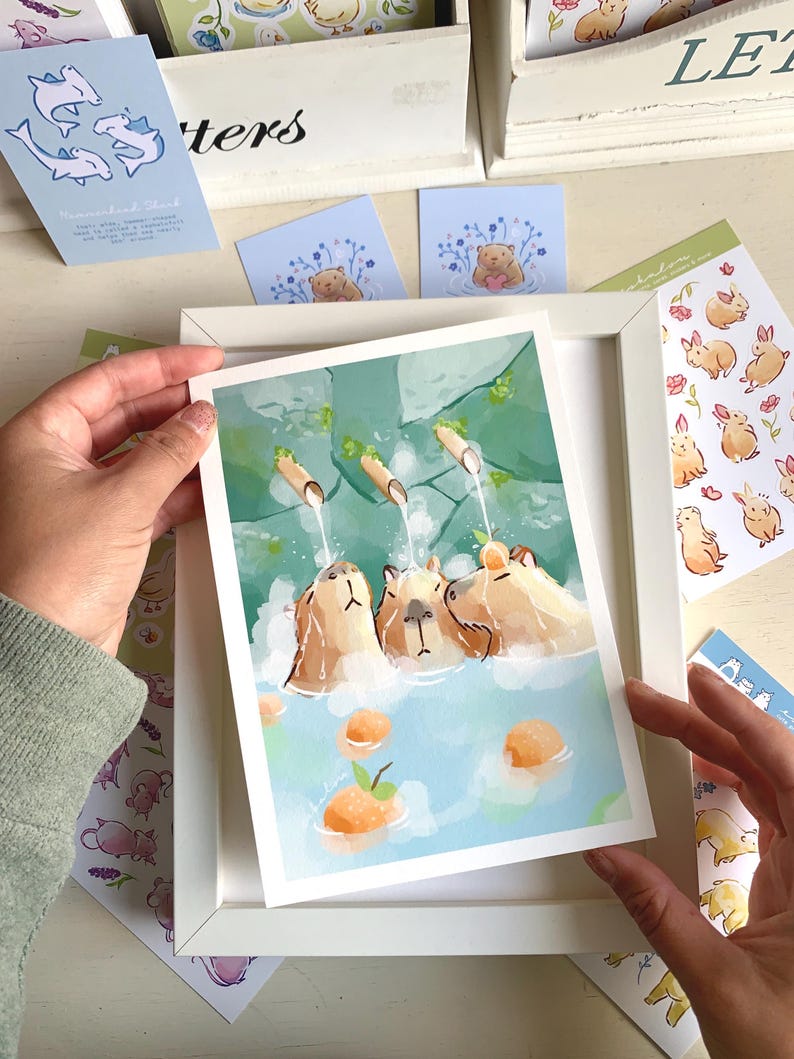 May include: A framed art print depicting capybaras in a hot spring. The watercolour illustration shows three capybaras in a blue pool with orange fruit. Other stationery items with animal illustrations are visible.