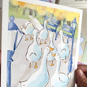 May include: Watercolor illustration of a flock of white geese in a park setting. The artwork features a soft color palette with blue, yellow, and green hues. The geese are depicted with detailed features, standing near a park bench and a lamppost.