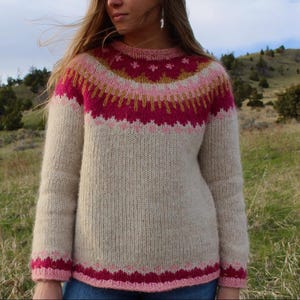 Könnte beinhalten: Ein cremefarbener Strickpullover mit einem pink-, gold- und burgunderfarbenen Muster am Joch. Der Pullover hat einen Rundhalsausschnitt und lange Ärmel.
