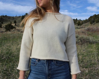 May include: A white cropped sweater with long sleeves and a ribbed texture. The sweater is worn by a person standing in a field.