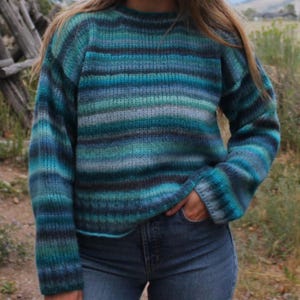 Könnte beinhalten: Ein gestreifter Pullover in Blautönen, Grün und Grau. Der Pullover hat einen Rundhalsausschnitt und lange Ärmel.