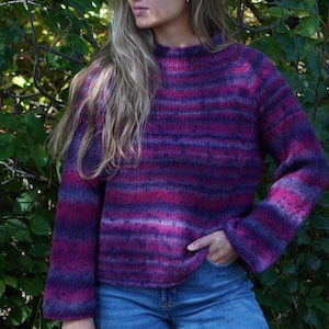 May include: A hand-knitted jumper with a mock neck and long sleeves. The jumper features horizontal stripes in shades of purple, pink, and blue. The garment is made of a soft, textured yarn, and is paired with blue jeans.