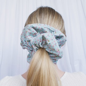 May include: A light blue scrunchie with a small floral pattern. The scrunchie is tied around a ponytail.