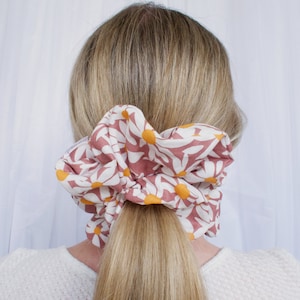 May include: A pink and white scrunchie with a daisy print. The scrunchie is tied around a ponytail.