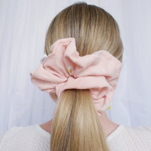 May include: A light pink scrunchie with a daisy pattern is wrapped around a blonde ponytail. The scrunchie is made of a soft, textured fabric. The background is a white curtain.