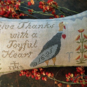 May include: A beige pillow with a cross-stitch design featuring a turkey and the text "Give Thanks with a Joyful Heart This is", a traditional Thanksgiving theme.