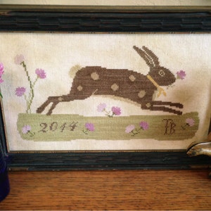 May include: A framed cross-stitch embroidery depicting a brown rabbit with white spots running through a field of pink flowers. The embroidery is dated "2014" and signed "TB".