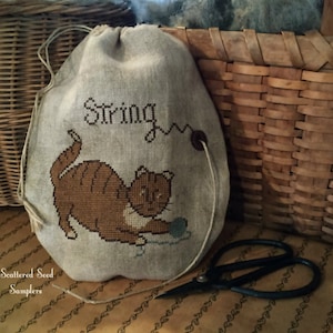 May include: A beige drawstring bag with a cat playing with a ball of yarn embroidered on it. The bag has the word "String" embroidered on it. The bag is sitting on a brown surface with a pair of black scissors.