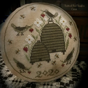 May include: A round, beige cross-stitch design featuring two beehives, birds, bees, flowers, and the year 2020. The design is framed by a stitched border. The text "Scattered Seed Samplers C2020" is in the upper right corner.