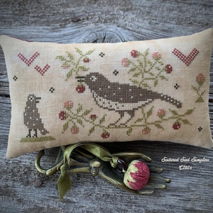 Puede incluir: Un cojín rectangular beige bordado a punto de cruz, con dos pájaros y elementos florales. Un pájaro está posado en una rama con bayas, el otro debajo. El diseño incluye acentos rojos y verdes, con el texto "Scattered Seed Samplers ©2024".