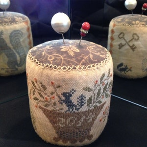 Puede incluir: Tres alfileteros redondos de tela con diferentes diseños bordados. Los alfileteros son beige con detalles marrones y presentan una ardilla, llaves y una cesta de bayas. Cada alfiletero tiene un alfiler de aspecto perlado en la parte superior.