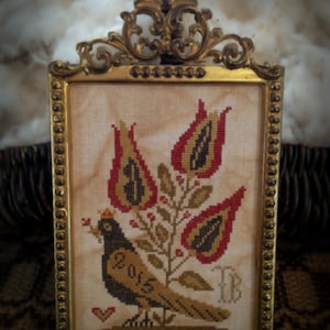 May include: A framed cross-stitch sampler featuring a stylized bird with a crown, surrounded by red and gold tulips. The sampler is signed "TB" in the lower right corner and "2015" on the bird's chest. The frame is gold with a beaded border.
