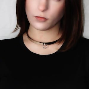 Puede incluir: Una mujer con el pelo castaño corto lleva un collar negro de gargantilla con un pequeño colgante circular. Lleva una camiseta negra de manga corta y tiene una expresión neutral en la cara.