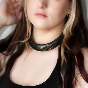 May include: A woman with long blonde hair wearing a black tank top and a black corset with silver buckles and laces. She is wearing a black choker necklace.