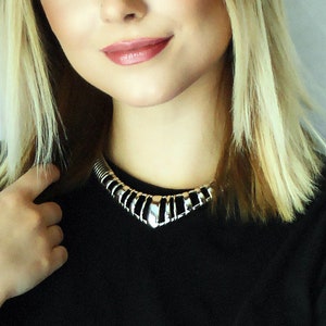 May include: A woman with long blonde hair wearing a black t-shirt and a silver necklace. She is looking at the camera and has her arms crossed.