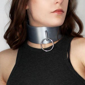 Puede incluir: Un collar gris con un detalle de anillo plateado. El collar es ancho y se coloca alrededor del cuello. La persona de la imagen lleva una camiseta sin mangas gris oscuro.