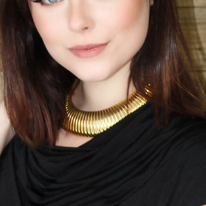 May include: A woman wearing a black top and a gold-colored collar necklace. The necklace is a thick, ribbed design that sits close to the neck. The top has a draped neckline and short sleeves. The background is a wooden wall.