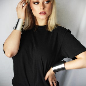 May include: A woman wearing a black t-shirt and black jeans with two silver metal cuffs on her wrists.