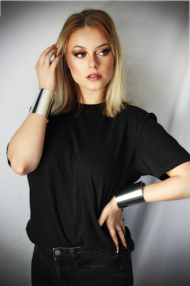 May include: A woman wearing a black t-shirt and black jeans with two silver metal cuffs on her wrists.
