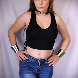 May include: A woman wearing a black crop top with a silver choker necklace and blue jeans. She is wearing silver bracelets on both wrists.