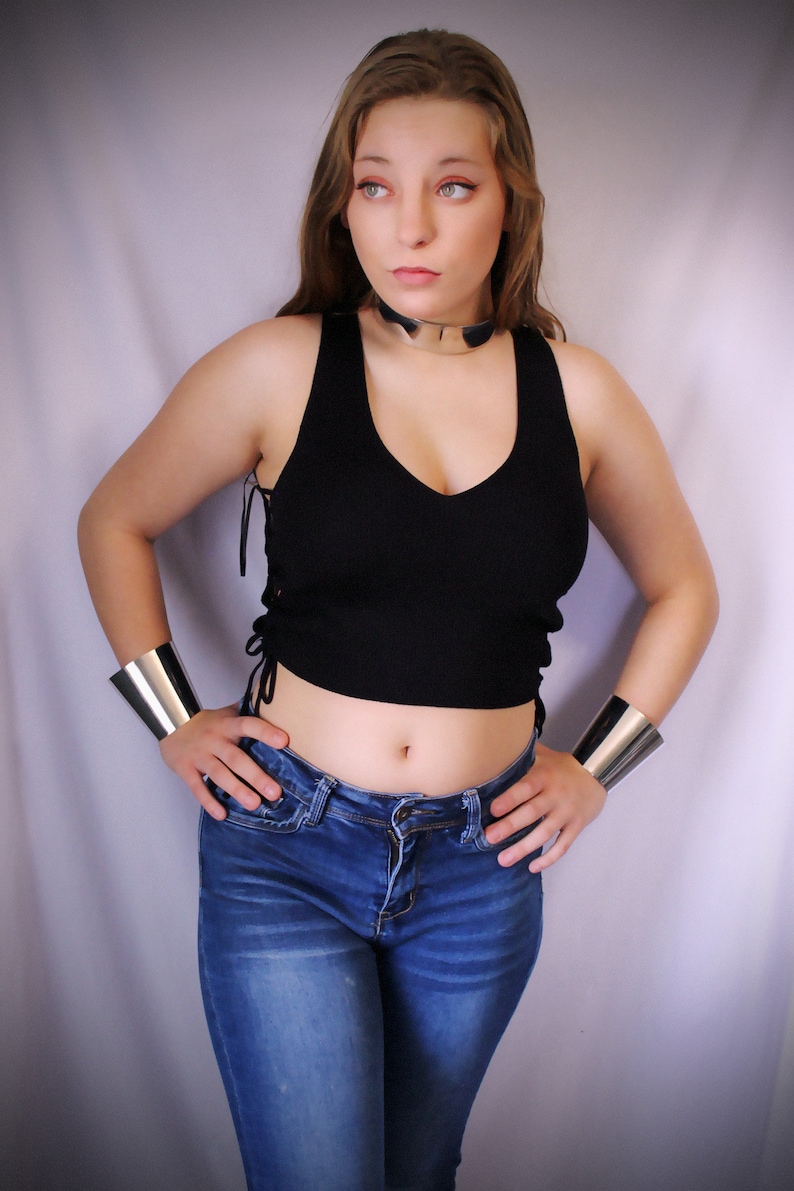 May include: A woman wearing a black crop top with a silver choker necklace and blue jeans. She is wearing silver bracelets on both wrists.