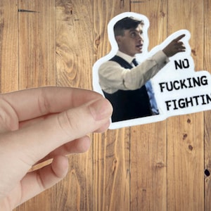 May include: A white sticker with the text "NO FUCKING FIGHTING" and an image of a man pointing. The sticker is held by a hand against a wooden background. The man is wearing a vest, white shirt, and tie.